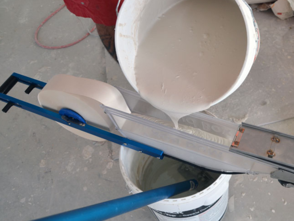 A drywall finisher fills the Mud Box Pro by pouring joint compound straight from the bucket to the mud chamber. This shows how easily it is to fill the Mud Box Pro as opposed to a drywall bazooka which requires a mud pump and gooseneck attachment.