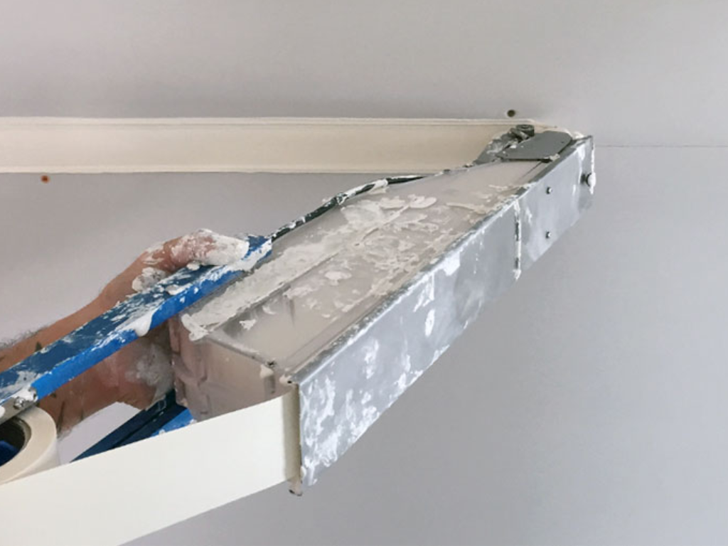 A drywall finisher uses the Mud Box Pro to simultaneously apply joint compound and paper tape to a horizontal drywall joint. The result is a clean tape run with very little mess.